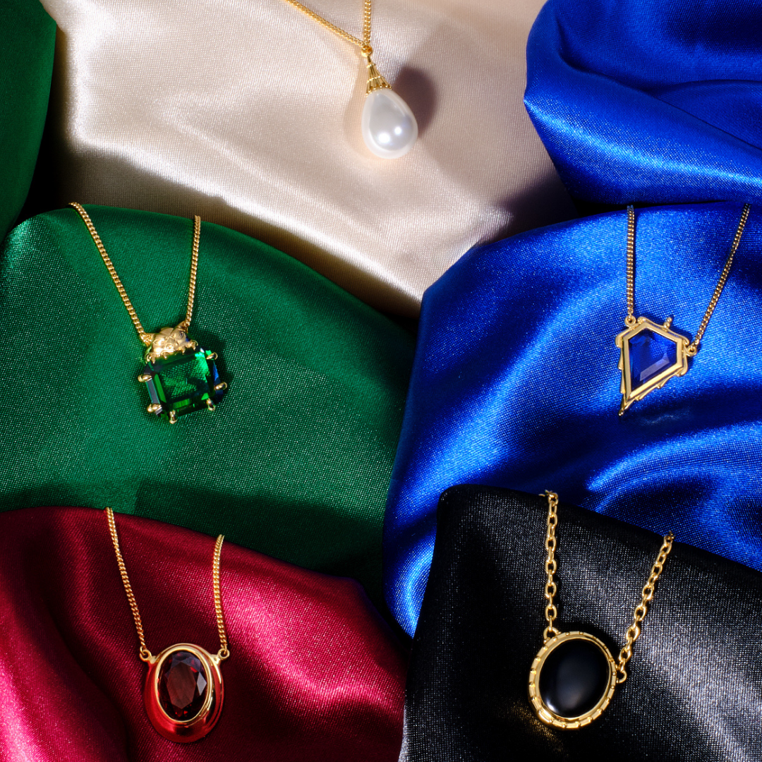 Magic: The Gathering X RockLove Mox Pearl, Sapphire, Jet, Ruby, and Emerald Necklaces — set in 14K gold
plated sterling silver, [context: Five gold pendant necklaces displayed on draped satin fabric in beige, green, blue, red, and black. The necklaces feature distinct pendants: a white pearl, a rectangular green gemstone, an oval red gemstone, and a geometric blue gemstone.]
