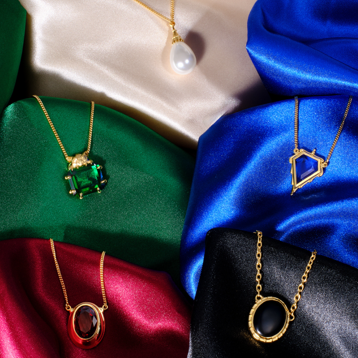 Magic: The Gathering X RockLove Mox Pearl, Sapphire, Jet, Ruby, and Emerald Necklaces — set in 14K gold
plated sterling silver, [context: Five gold pendant necklaces displayed on draped satin fabric in beige, green, blue, red, and black. The necklaces feature distinct pendants: a white pearl, a rectangular green gemstone, an oval red gemstone, and a geometric blue gemstone.]