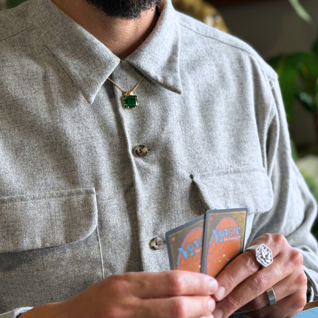 Magic: The Gathering X RockLove Mox Emerald Necklace —  Emerald set in 14K gold plated sterling silver, [context: a close-up lifestyle shot of a man wearing a light gray textured button-down shirt, a gold necklace with a green square pendant, and silver rings, holding two Magic: The Gathering cards.]