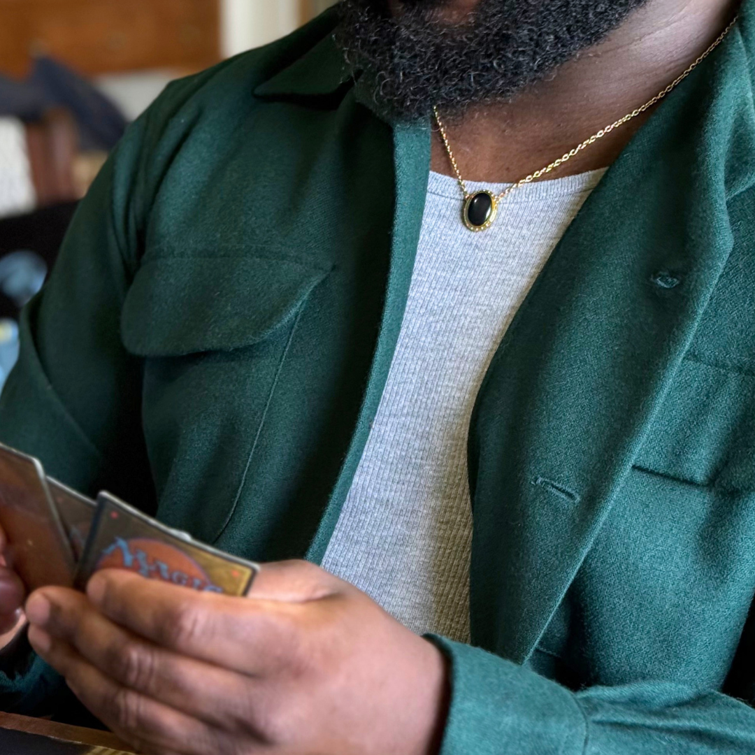 Magic: The Gathering X RockLove Mox Jet Necklace — Onyx set in 14K gold
plated sterling silver, [context: a close-up lifestyle shot of a man wearing a dark green wool-blend blazer over a light gray ribbed t-shirt, accessorized with a gold chain necklace featuring a black oval pendant. He is holding Magic the Gathering cards in his hands.]