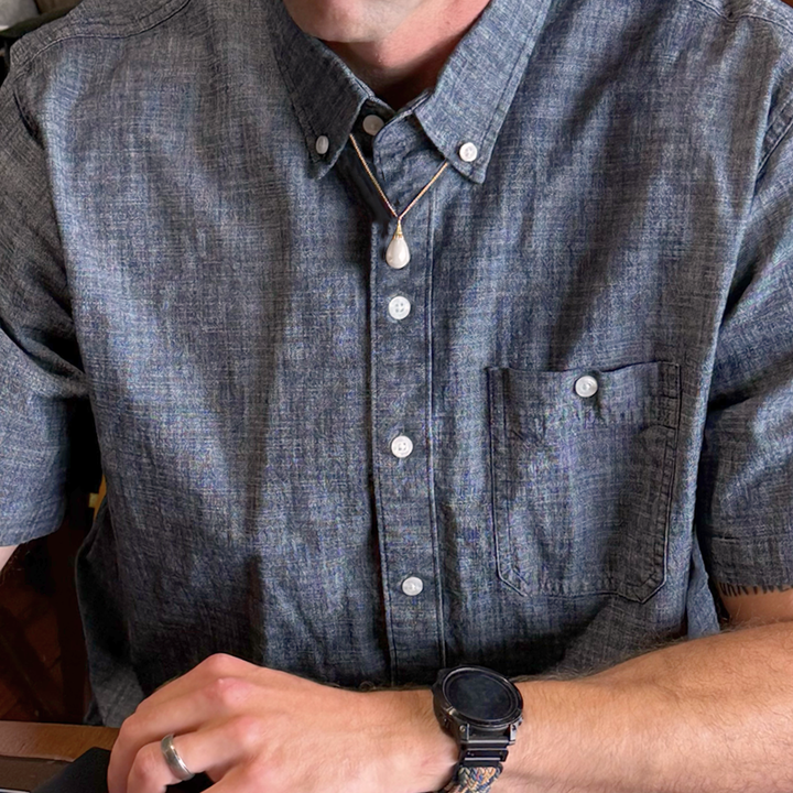 Magic: The Gathering X RockLove Mox Pearl Necklace —  Pearl set in 14K gold
plated sterling silver, [context: a close-up of a man wearing a textured slate blue short-sleeve button-down shirt with white buttons, a white teardrop pendant necklace, and a black smartwatch.]