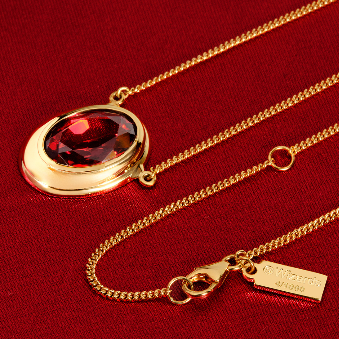 Magic: The Gathering X RockLove Mox Ruby Necklace —  Ruby set in 14K gold
plated sterling silver, [context: gold chain necklace with an oval gold locket pendant featuring a faceted red gemstone, displayed on a textured red fabric background. The tag on the clasp reads '4/1000'.]