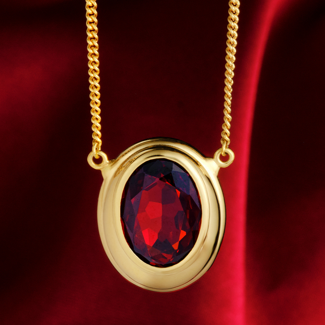 Magic: The Gathering X RockLove Mox Ruby Necklace —  Ruby set in 14K gold
plated sterling silver, [context: a gold oval pendant necklace featuring a faceted deep red gemstone, suspended from a gold chain against a rich red satin background.]
