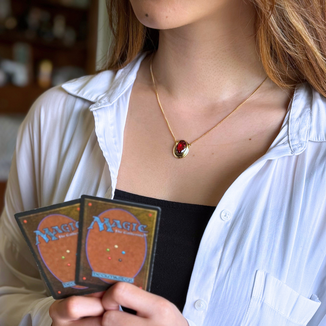 Magic: The Gathering X RockLove Mox Ruby Necklace —  Ruby set in 14K gold
plated sterling silver, [context: a gold necklace with a red oval pendant is worn by a woman in a white button-up shirt, holding two Magic: The Gathering cards.]