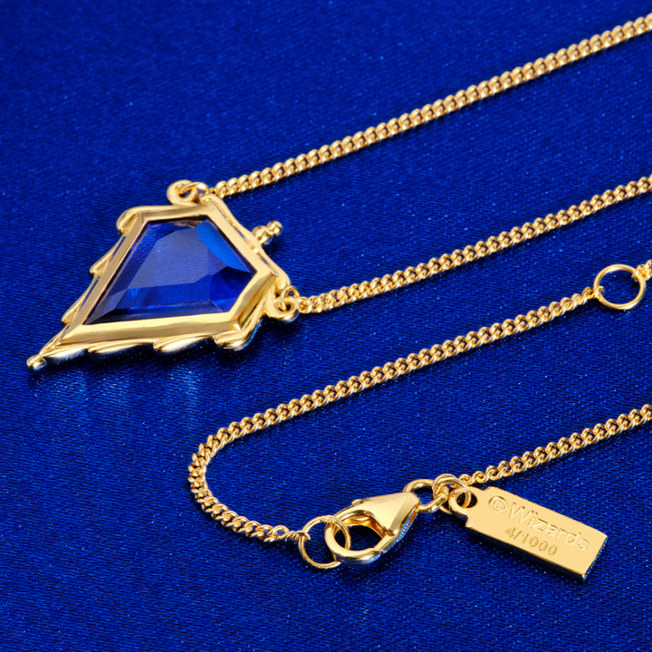 Magic: The Gathering X RockLove Mox Sapphire Necklace —  Sapphire set in 14K gold plated sterling silver, [context: a gold chain necklace with a geometric triangular pendant featuring a faceted blue gemstone, displayed on a textured blue background. The clasp end is shown with a brand tag engraved '4/1000'.]
