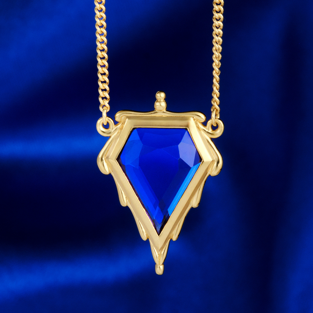 Magic: The Gathering X RockLove Mox Sapphire Necklace —  Sapphire set in 14K gold plated sterling silver, [context: a gold pendant necklace featuring a faceted blue gemstone in a shield-like frame, suspended from a gold chain against a deep blue satin background.]