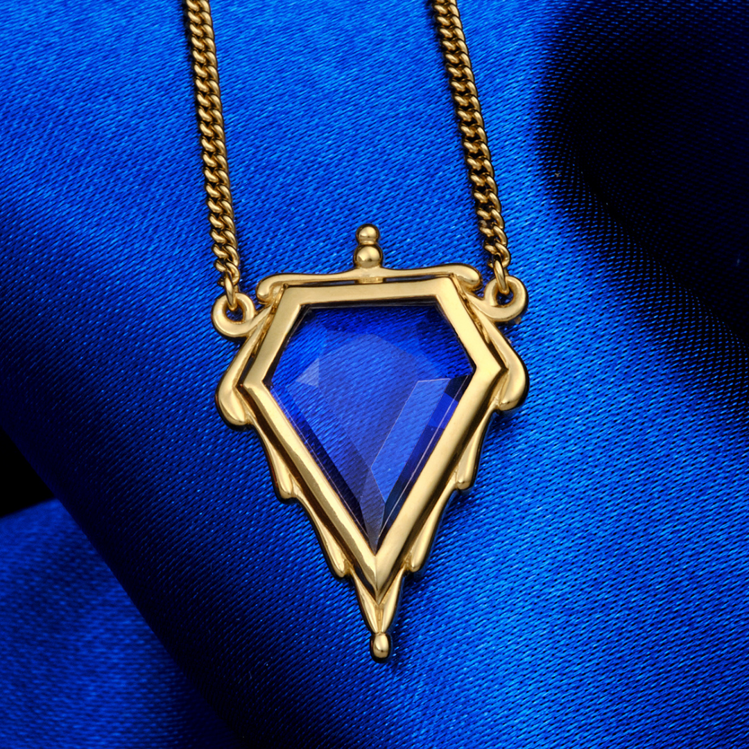 Magic: The Gathering X RockLove Mox Sapphire Necklace —  Sapphire set in 14K gold plated sterling silver, [context: a gold pendant necklace with a faceted blue gemstone, displayed on a textured royal blue fabric background.]
