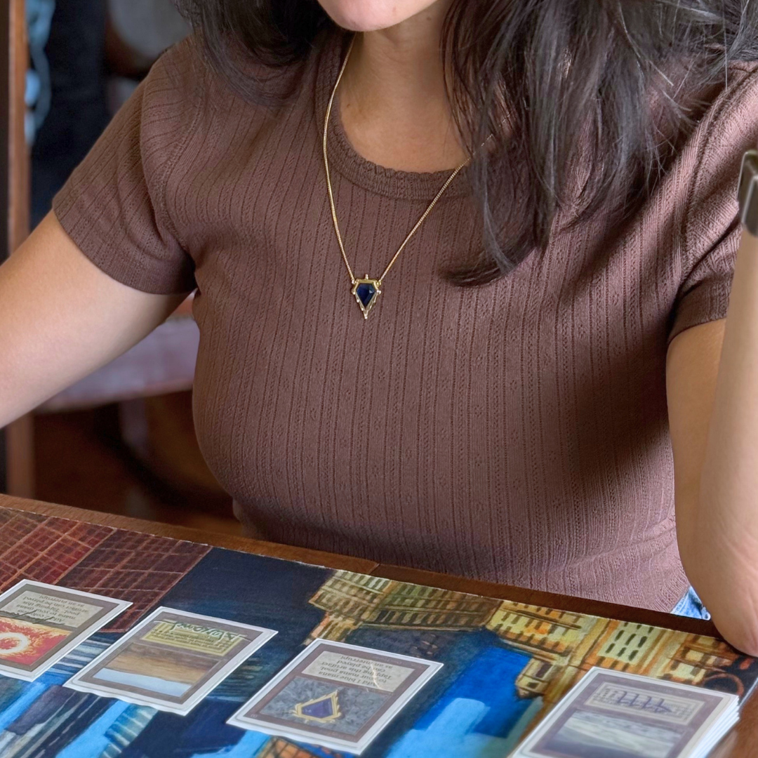 Magic: The Gathering X RockLove Mox Sapphire Necklace —  Sapphire set in 14K gold plated sterling silver, [context:  A woman wearing a brown ribbed short-sleeve top and a gold necklace with a dark blue pendant, seated at a table with Magic the Gathering cards in a game.]